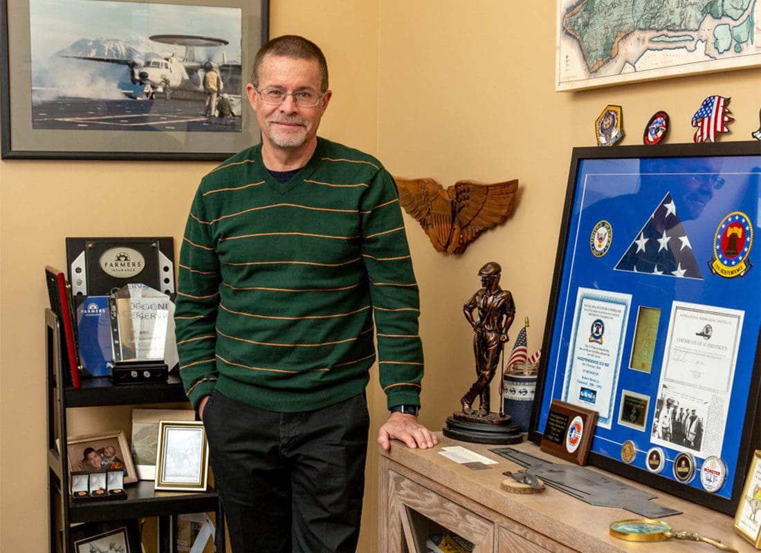 About Our Agency - Smiling Portrait of Patriotic Insurance Group Agency Owner and Veteran Rob Standing in his Office Next to a Display of Patriotic Badges and Certificates