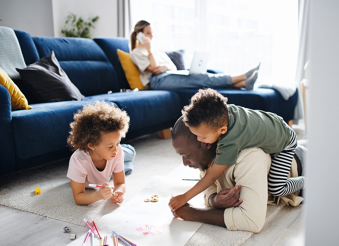 Personal Insurance - View of a Young Father Playing with His Two Young Children as They Draw on a Piece of Paper Spread Out on the Floor in the Living Room