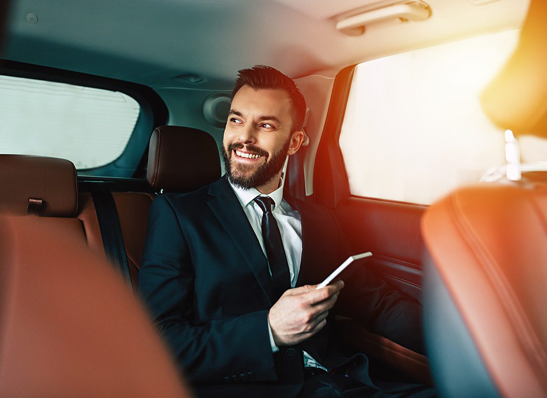 Service Center - Portrait of a Cheerful Young Businessman Sitting in the Back of a Car with Red Seats While Holding a Phone in his Hands